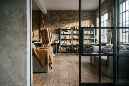 Atelier d'artiste converti en loft avec cloison vitrée coulissante en acier noir séparant espace de travail et salon, sol en chêne brut et mur en béton