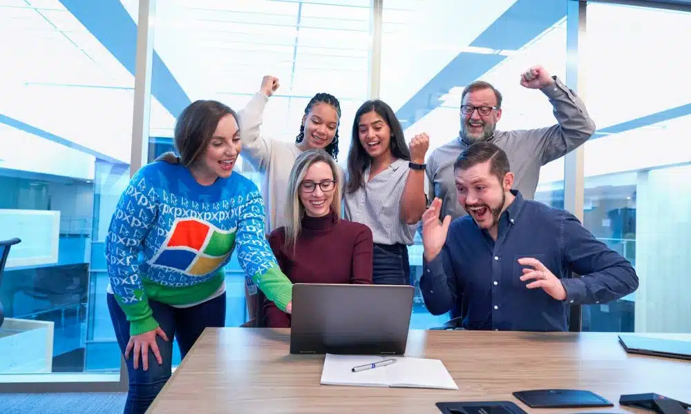 men and women sitting and standing by the table looking happy while staring at laptop