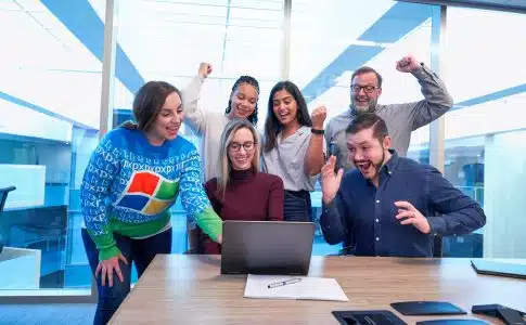 men and women sitting and standing by the table looking happy while staring at laptop